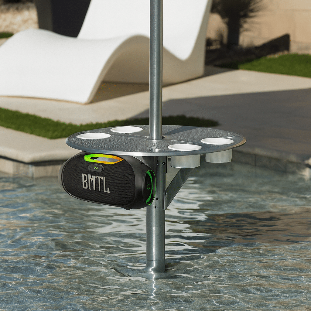 Water resistant 100W portable speaker mounted under a BajaMate pool table, shown in a resort-style backyard setup with clear water and modern outdoor furniture.