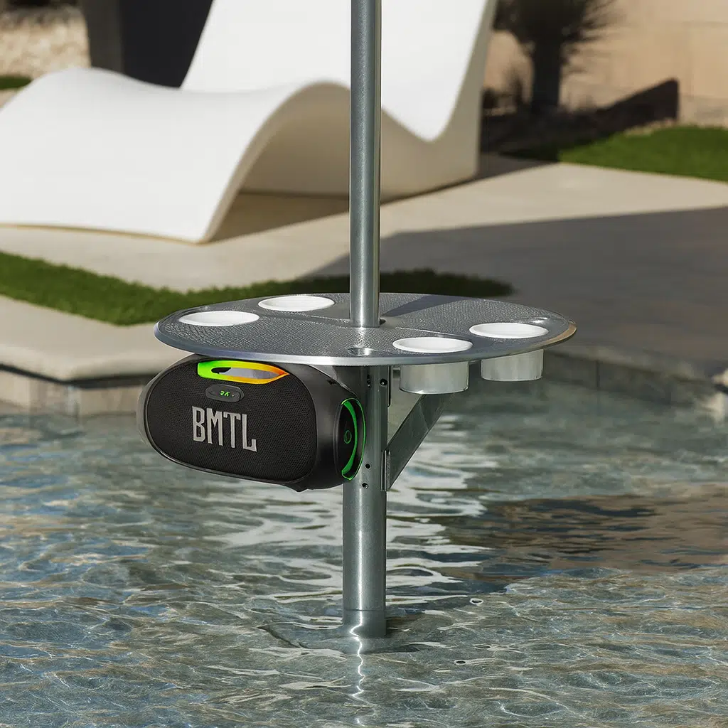 Water resistant 100W portable speaker mounted under a BajaMate pool table, shown in a resort-style backyard setup with clear water and modern outdoor furniture.